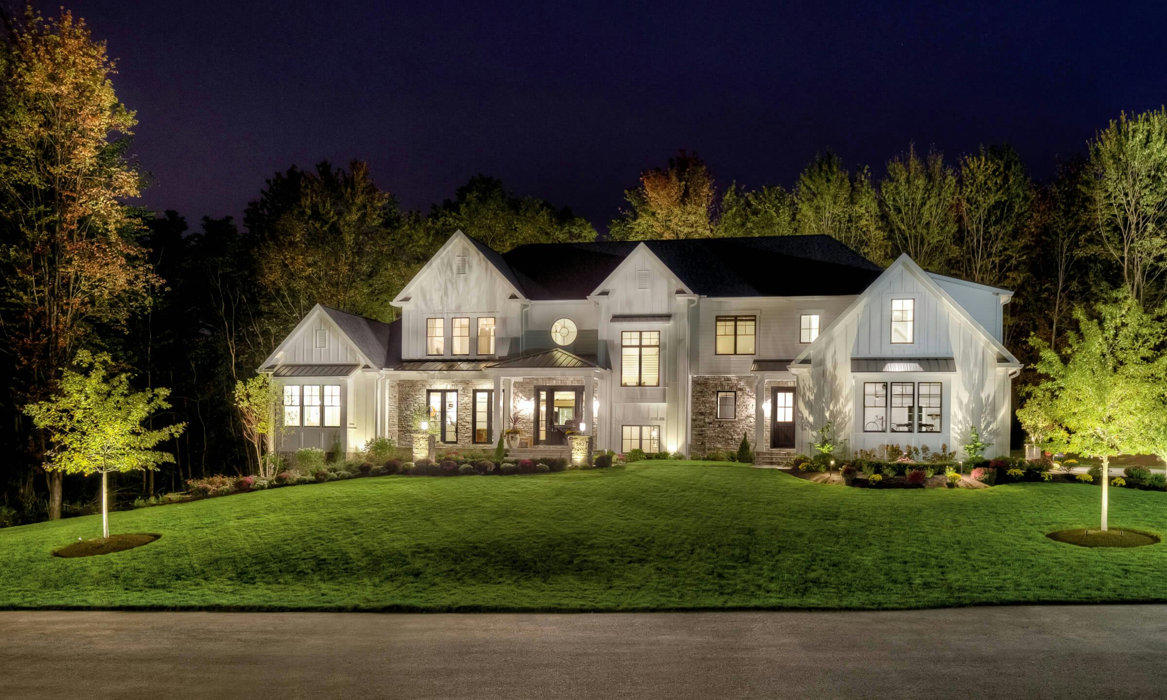 Exterior image of the front a modern farm house lit with landscape lighting, tree lights, and exterior lights at night 