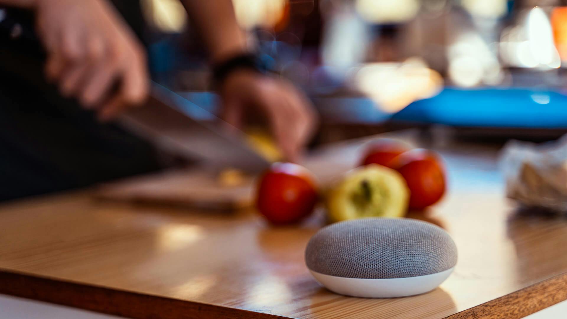 A grey Google Home sitting on a cutting board while someone slices tomatoes in the background