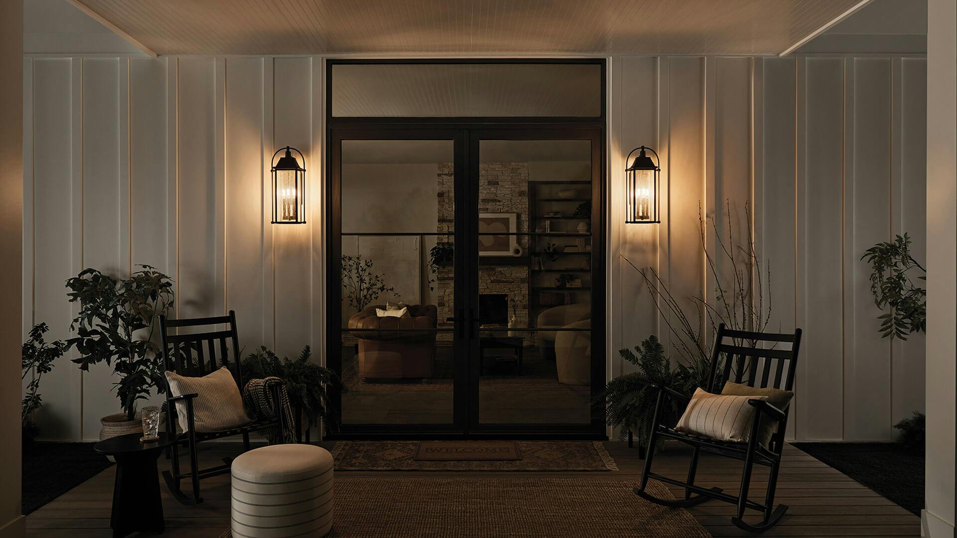 Covered patio with two rocking chairs and glass entry door flanked by two Harbor Row wall lights at night
