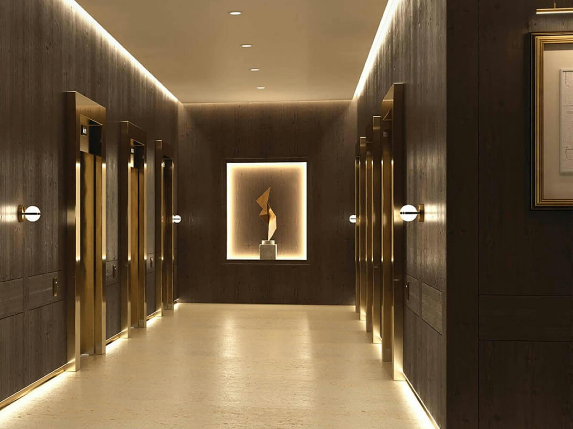 Interior corridor lit with variety of ceiling lights and sconces, lined by elevators