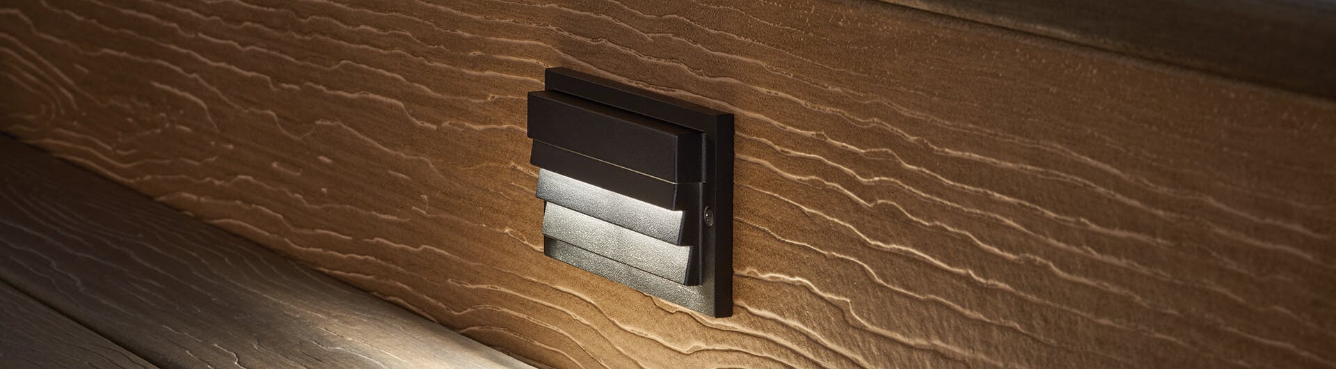 Close-up of a Louvered Step Light in Centennial Brass mounted against siding