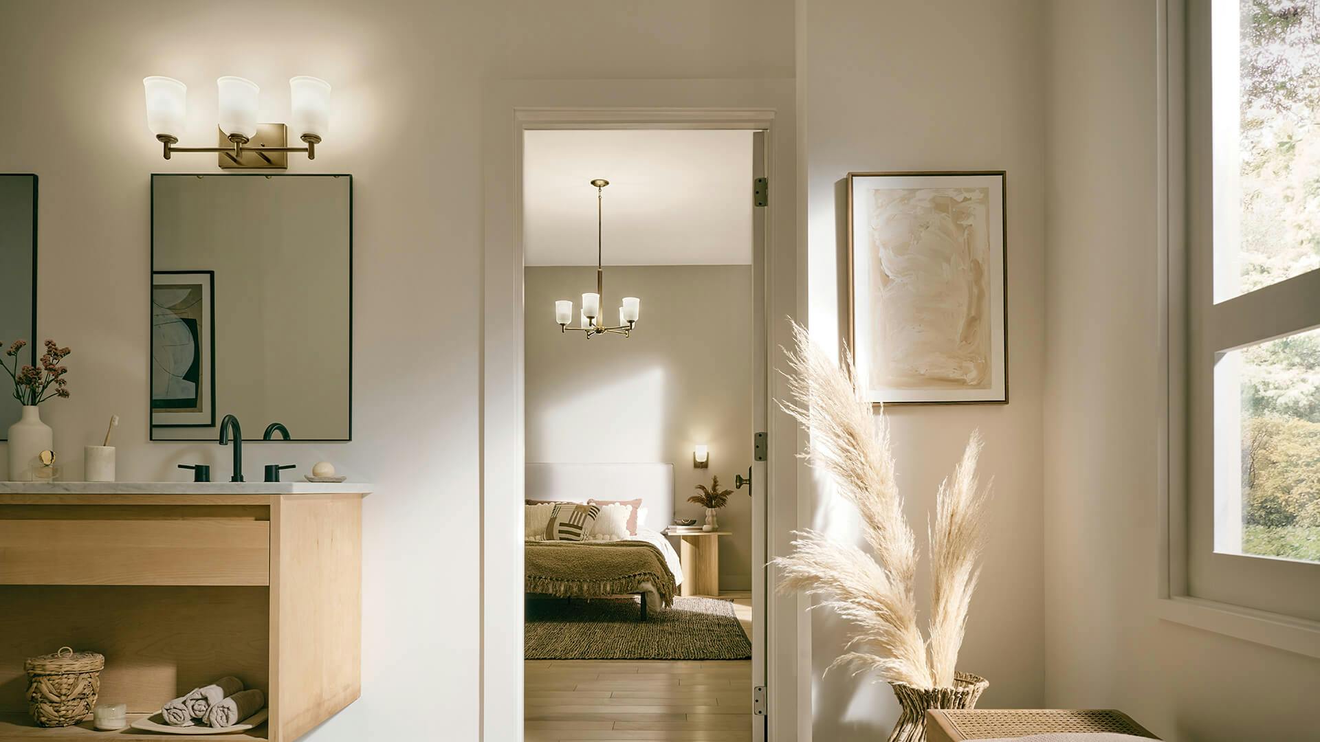 Bathroom looking into a doorway to a bedroom, warm cream and tan accents throughout featuring Shailene wall sconce above the vanity and chandelier in bedroom