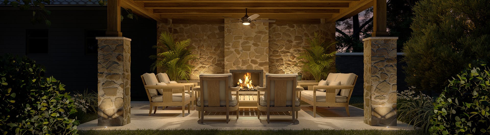 Outside patio at night with ceiling fan light and uplight along the back wall with a fireplace.
