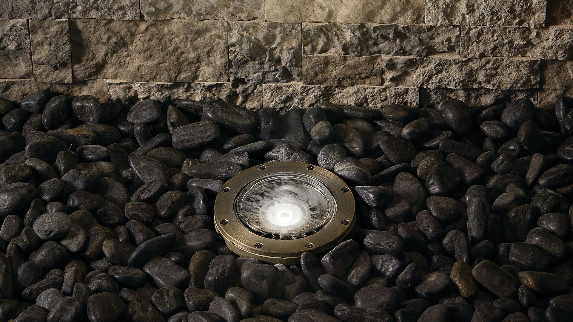 Close up of an in-ground light in gravel in front of a stone wall