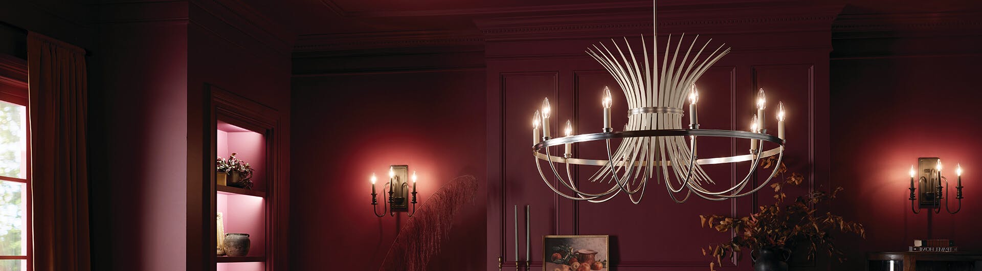 Dark red dining room lit by a brushed nickel Baile chandelier over the table with two sconces in the background