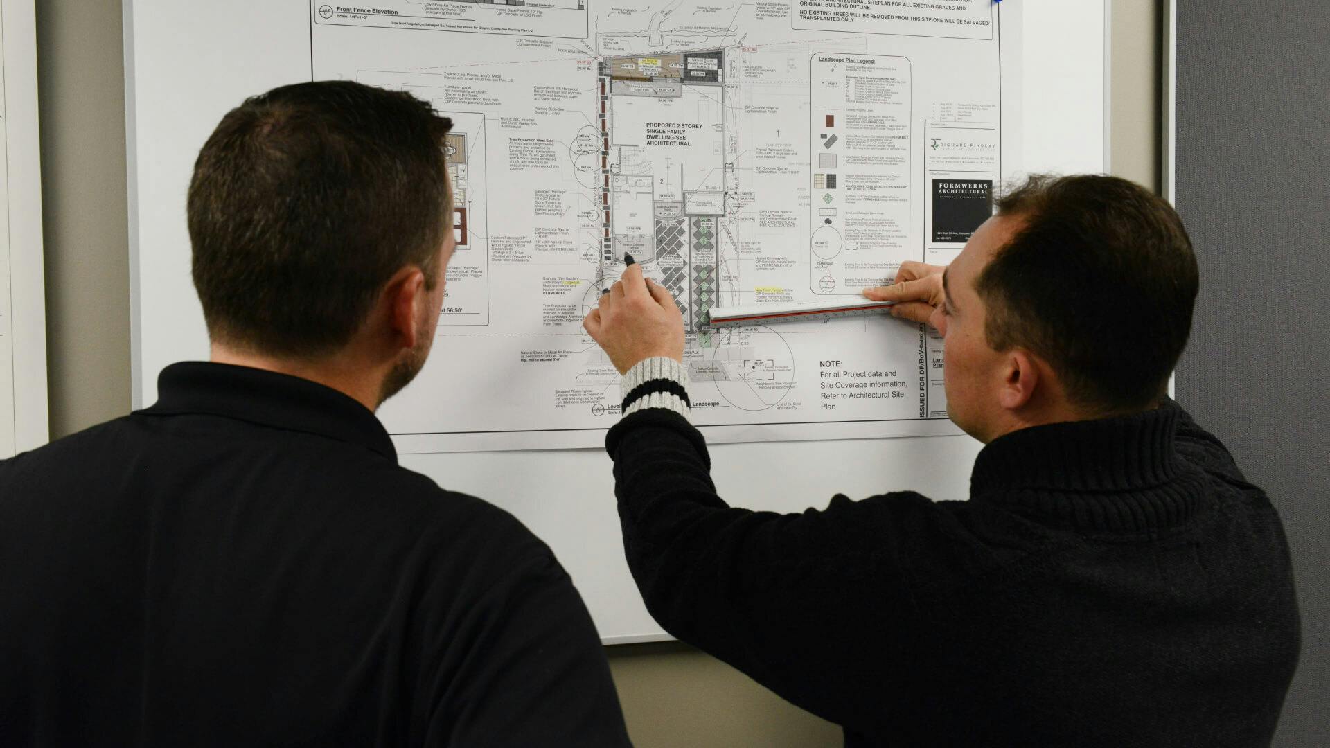 Two men standing in front of a blueprint on a wall, discussing.