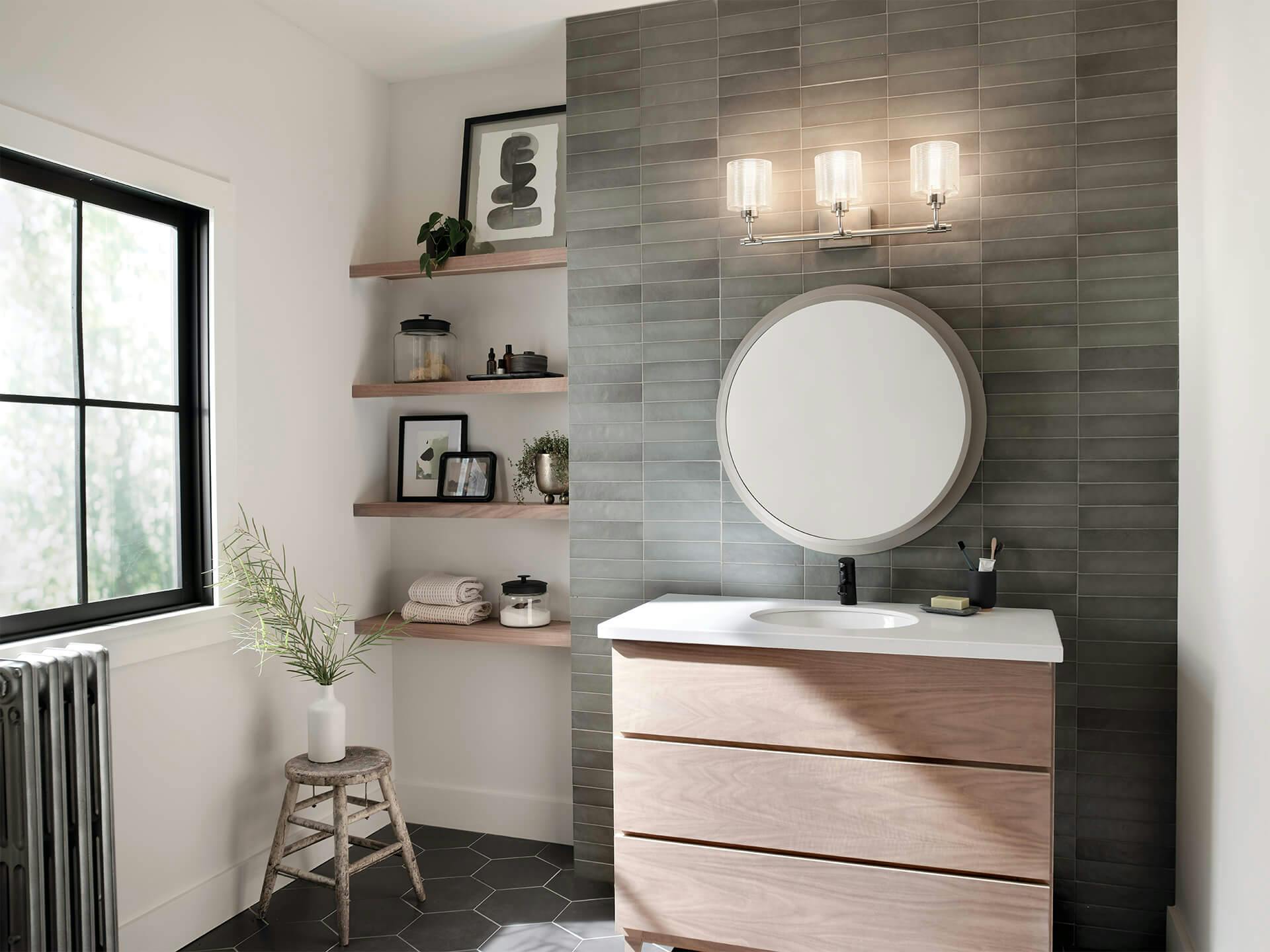 A modern bathroom with a Harvan vanity light installed on a grey brick textured wall above a mirror in the daytime with the lights turned on 
