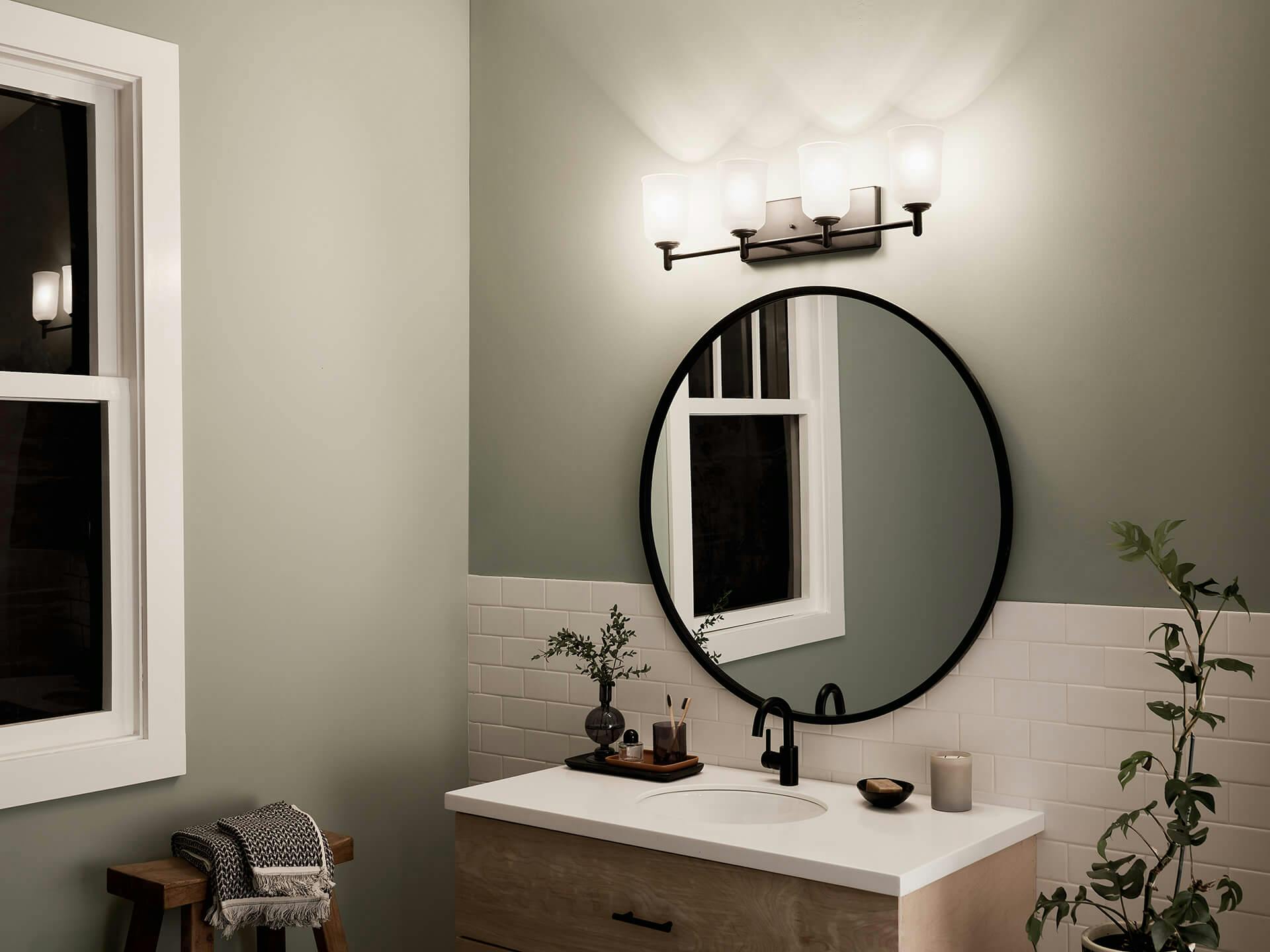 Corner of a bathroom vanity at night with a Shailene vanity light above a round mirror in black finish