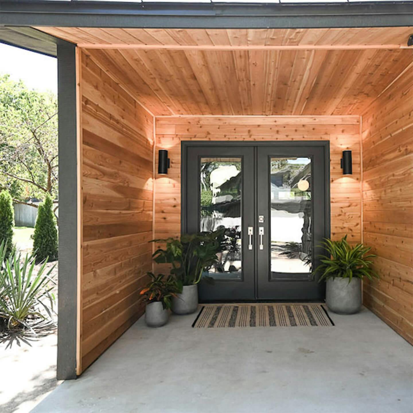 Exterior image of a modern front entrand with exposed wood siding with exterior wall lights mounted on either side of a black double door 