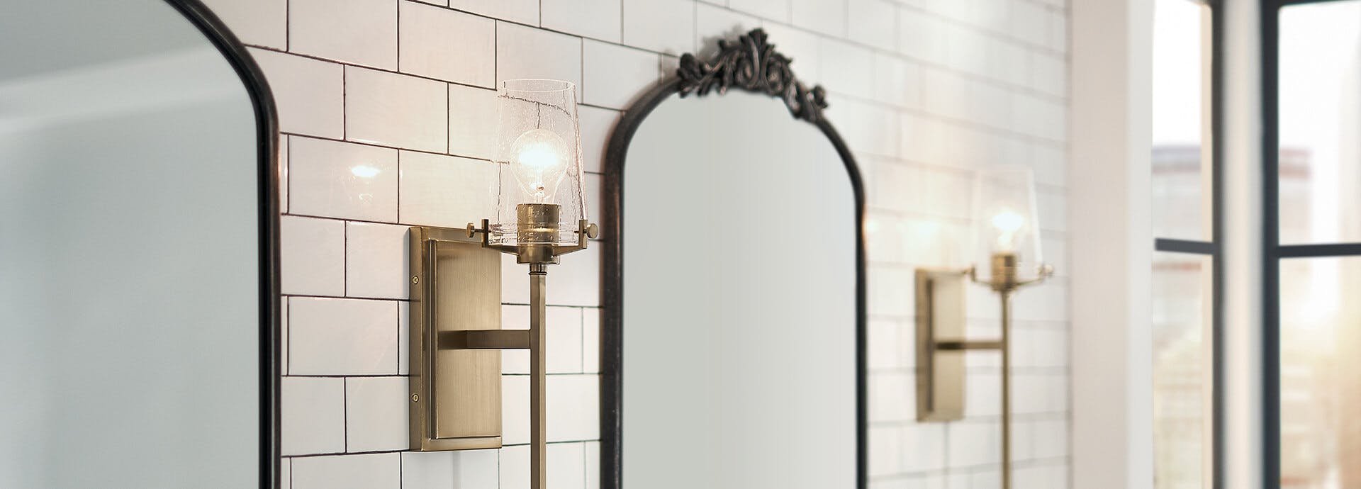 Two brass alton sconces on either side of two mirrors in a white tiled bathroom during the day