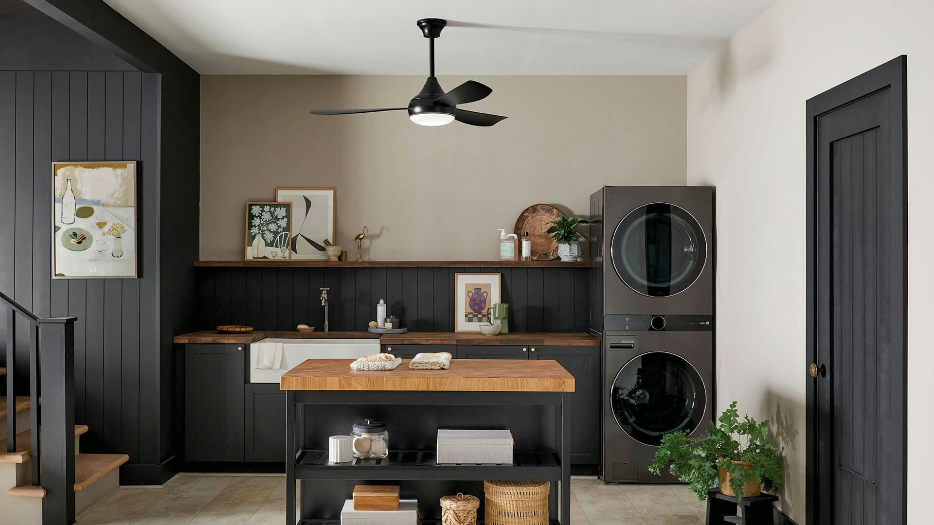 A downstairs laundry room with dark paneled walls featuring a black ample ceiling fan in the center.