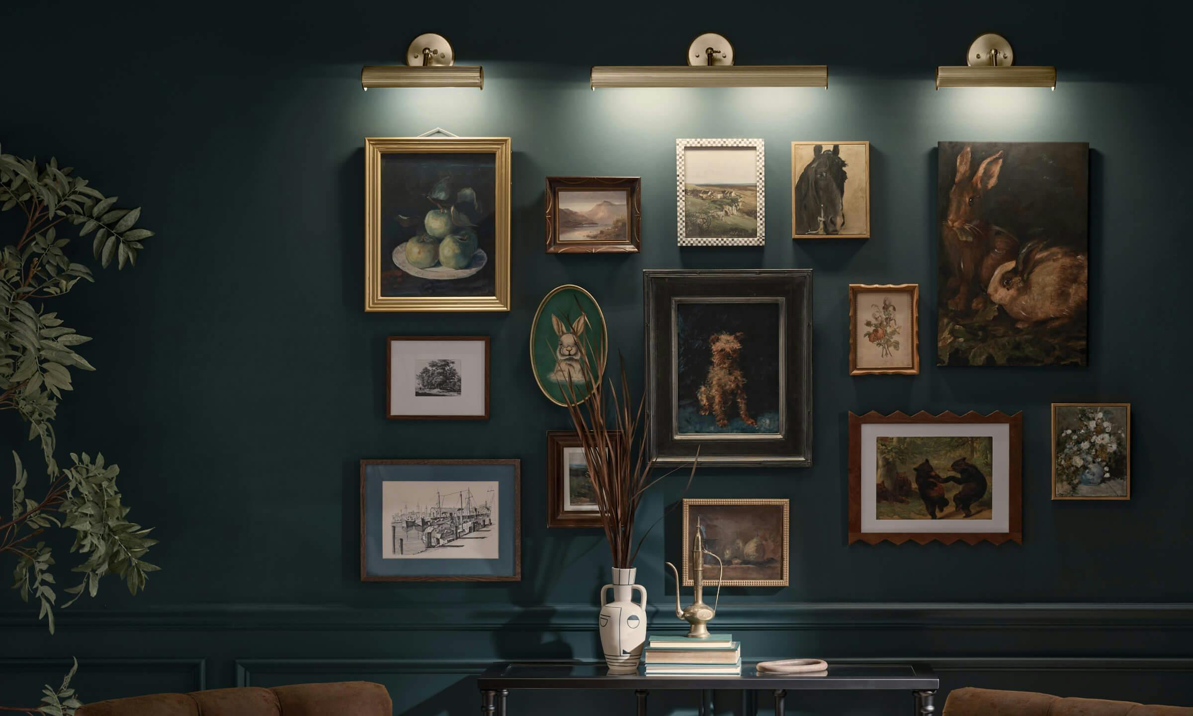 Den wall painted deep green with an arrangement of numerous pictures of varying sizes lit by three Midi sconces