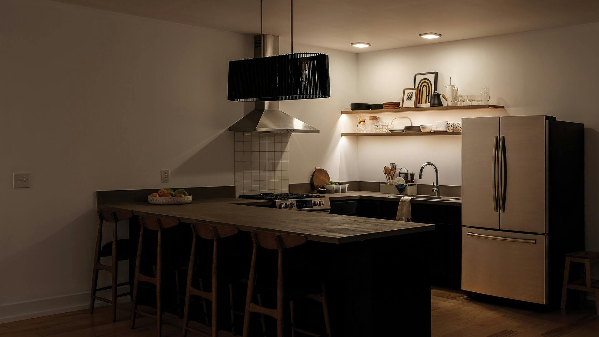 Kitchen in low-light with a Linara chandelier over the island while two Zeo and channel lights illuminate the back wall above the sink