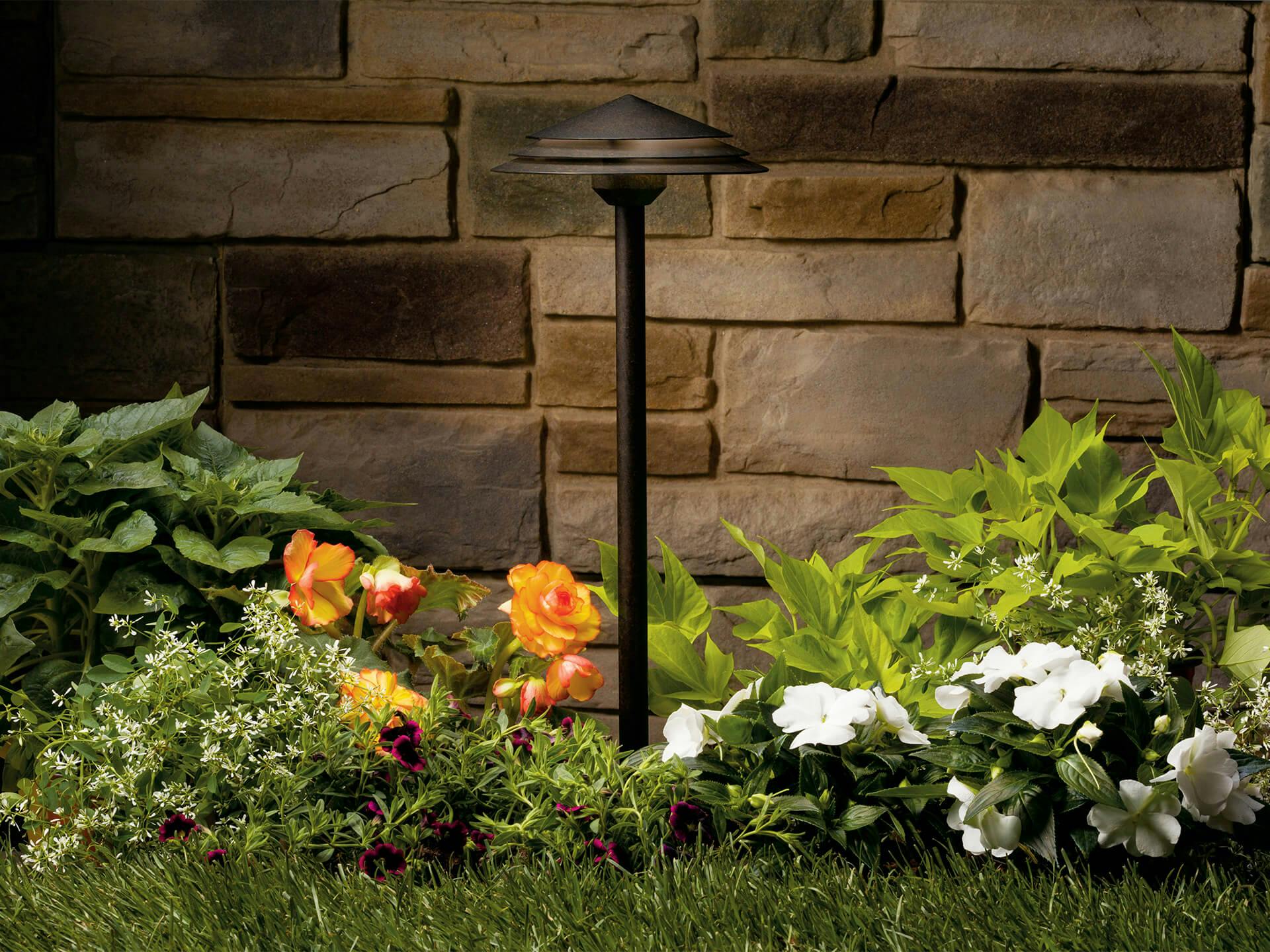 Close up of a path light in a garden with a stone wall behind it and flowerbed around it