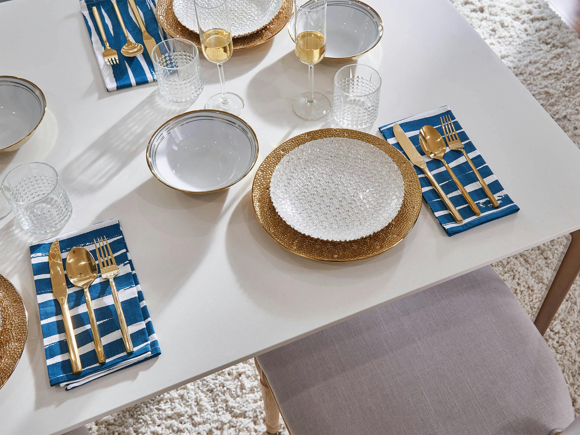 Lifestyle image of a white dinning room table top that has gold finished silverware and dishes for a dinner