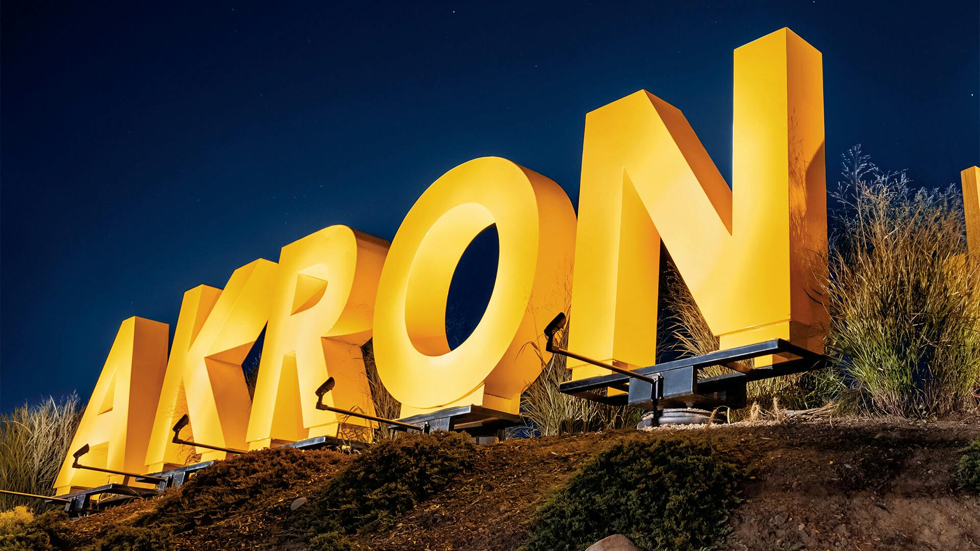 Close up on "Akron" part of the Akron Zoo sign, bright yellow letters with lights shining on each letter at night