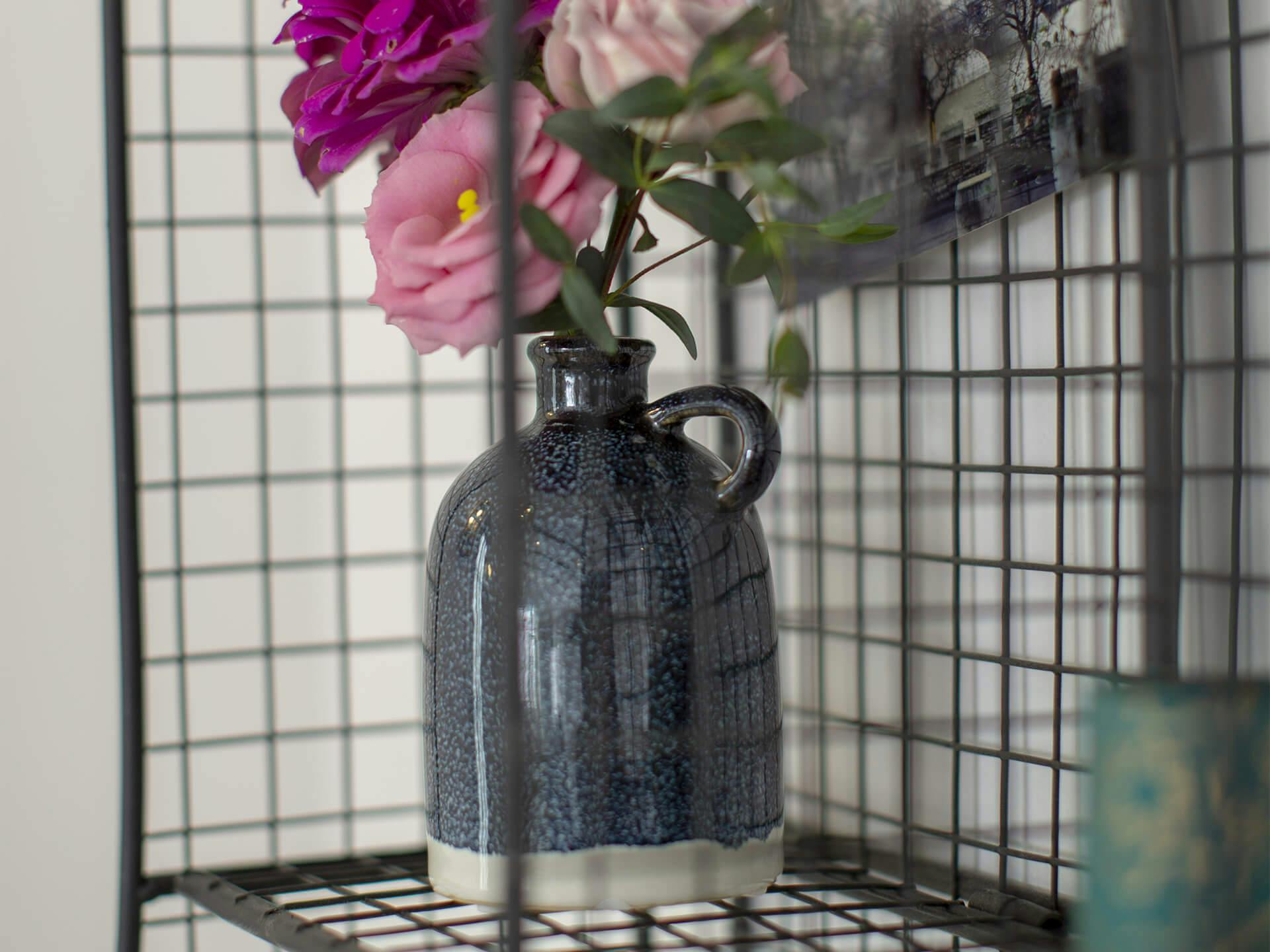 Lifestyle image of a grey jug vase filled with pink and violet flowers on a shelf