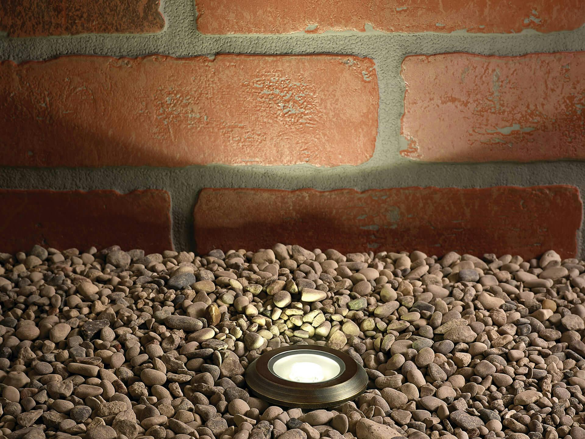 Close-up of a lit in-ground LED landscape light in centennial brass surrounded by gravel while lighting a brick wall