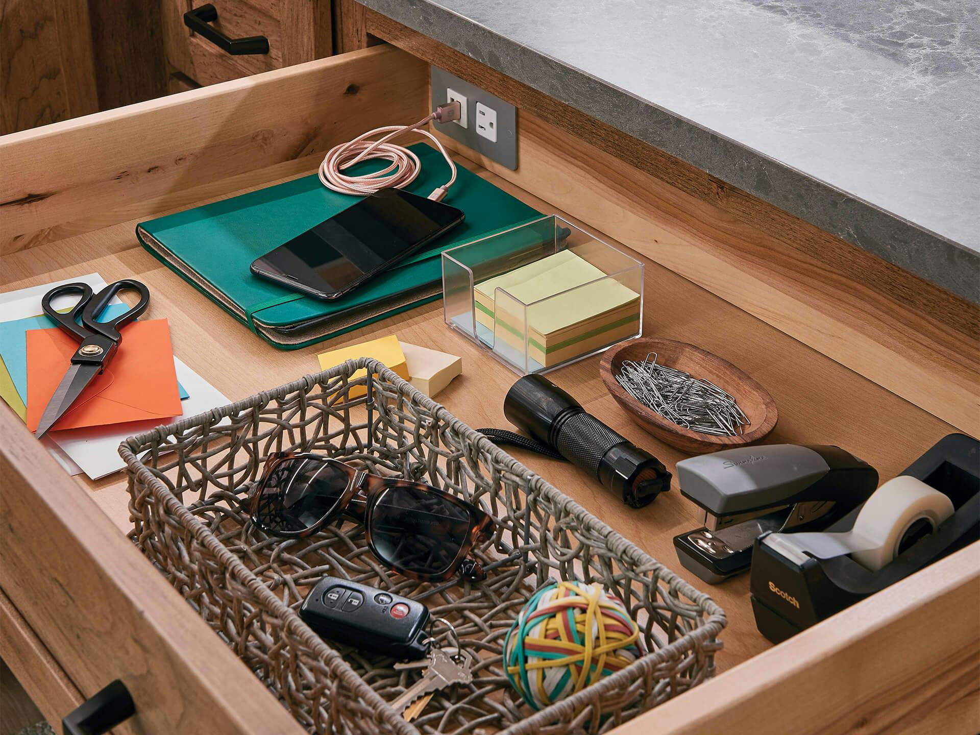 Open drawer containing a charging phone, keys, sunglasses and miscellaneous office supplies