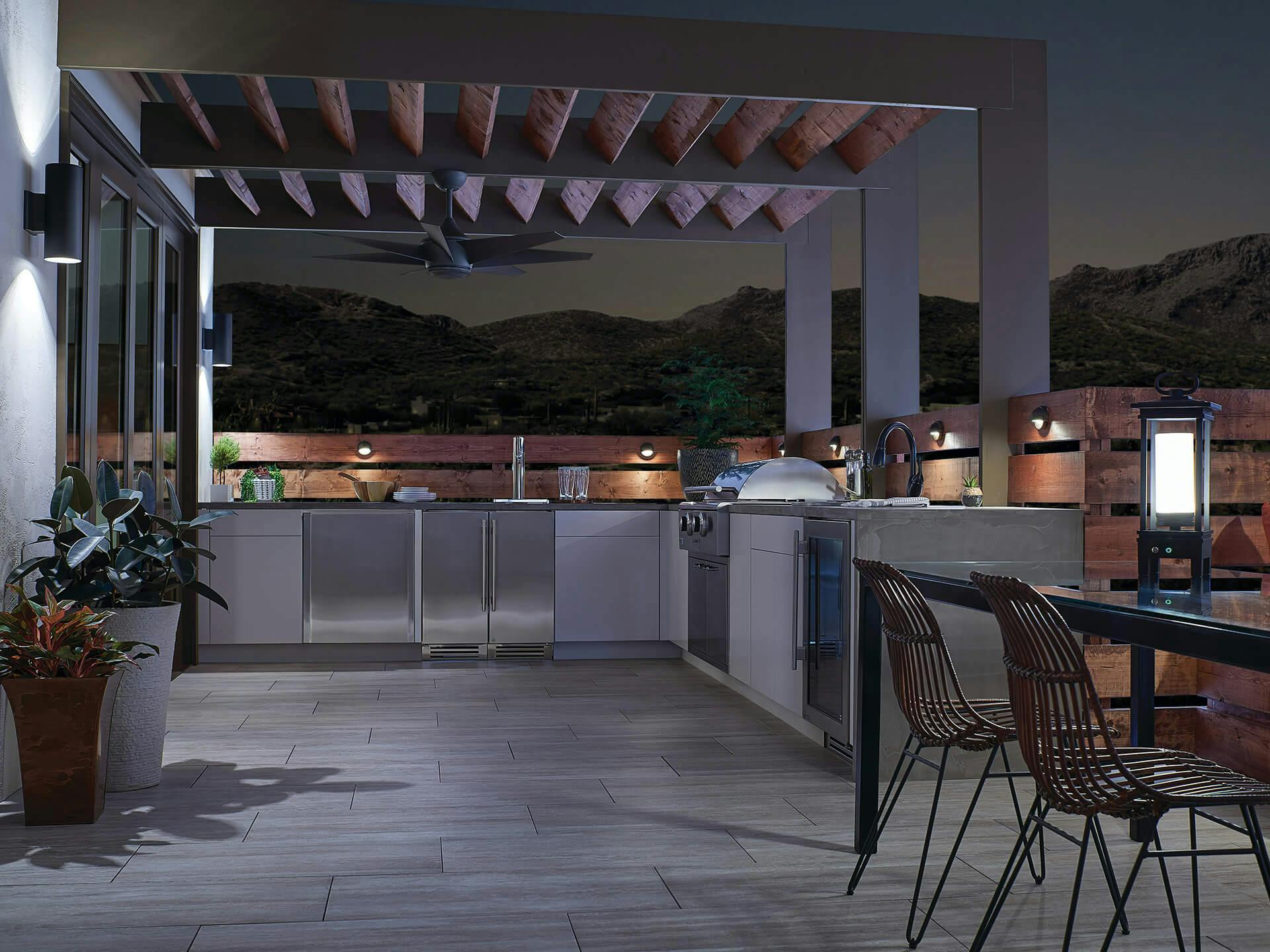 Spacious outdoor kitchen with Lehr 2 ceiling fan attached to a wooden overhang at night with accent lighting throughout