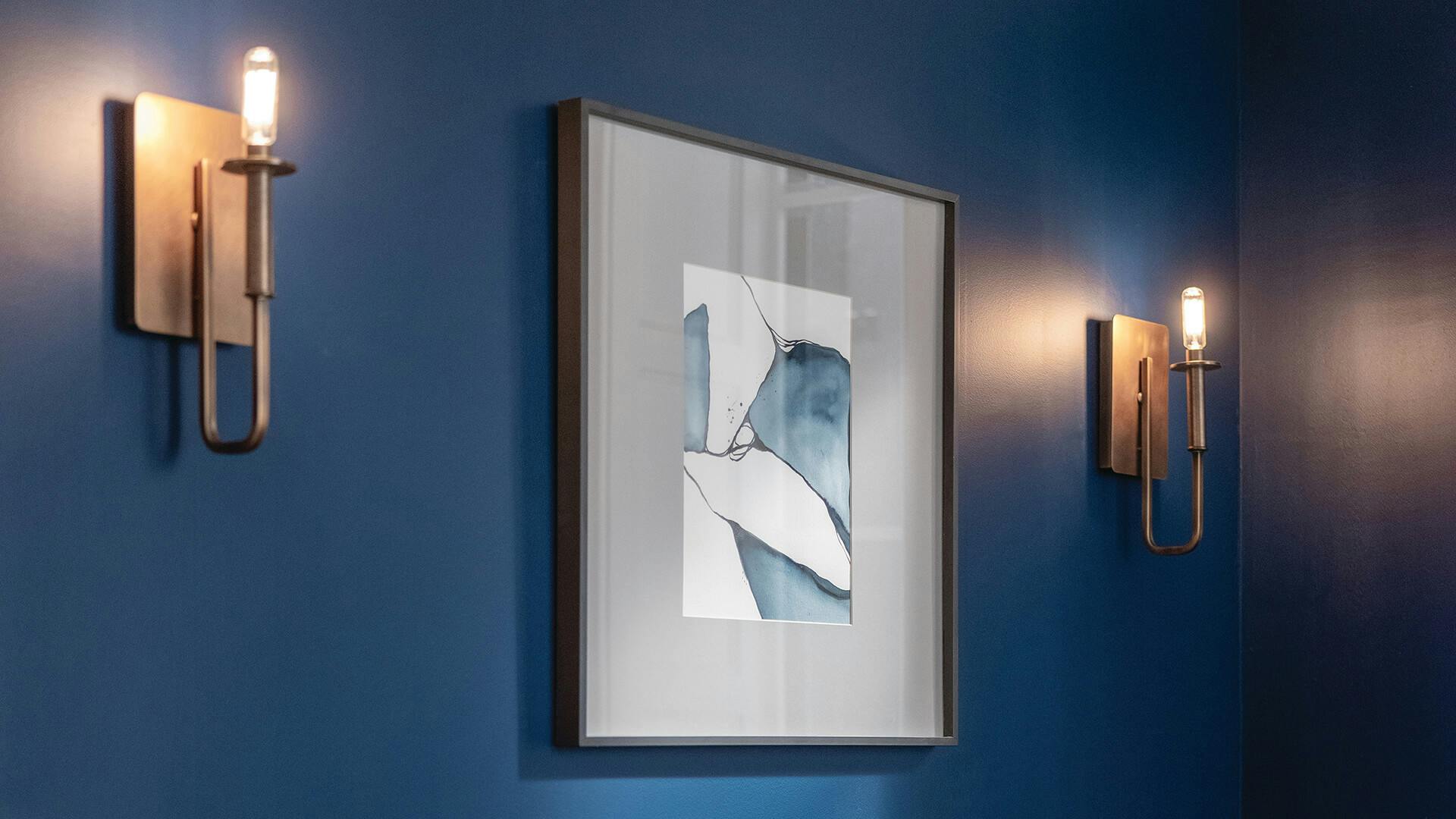Two lit Alden sconces against a deep blue wall surround a hung illustration