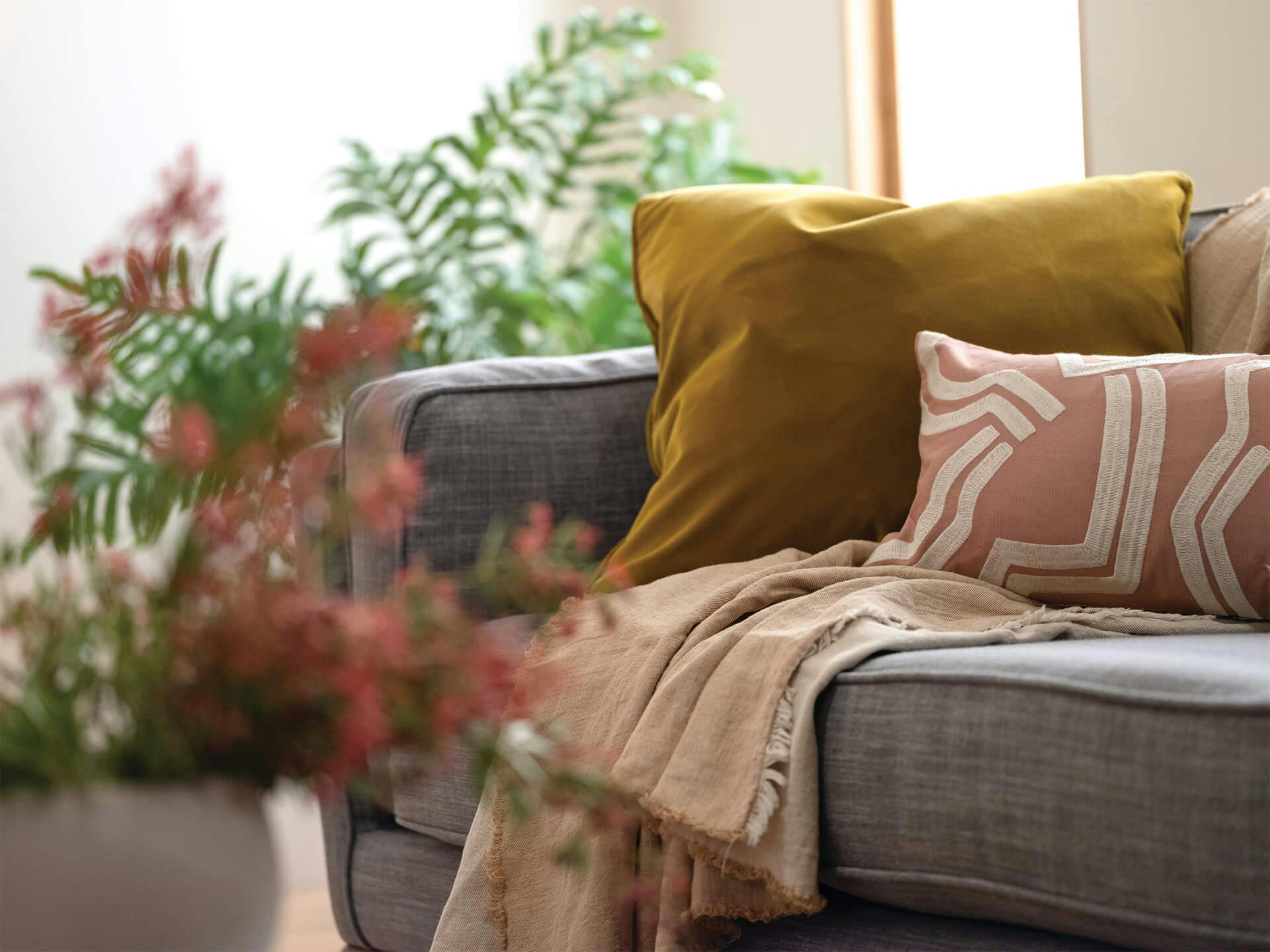 Lifestyle image of a grey couch with yellow and pinks pillows and a blanket with a vase full of wildflowers next to it 