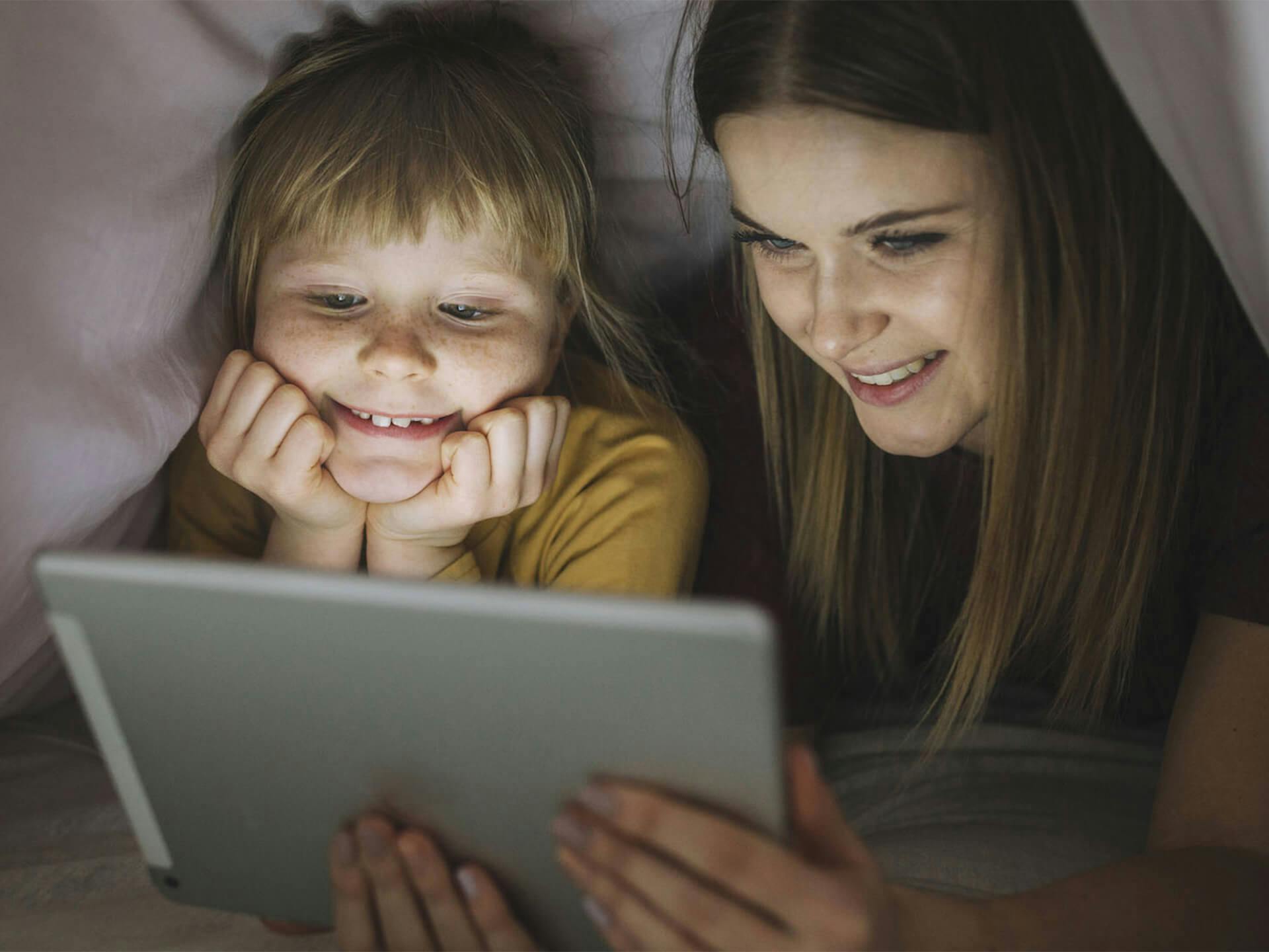 Lifestyle image of a mom and child looking at an Ipad underneath the blankets