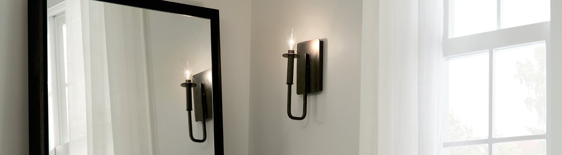Close up of a black finish Alden wall sconce in a corner of a white room with a mirror and window next to it