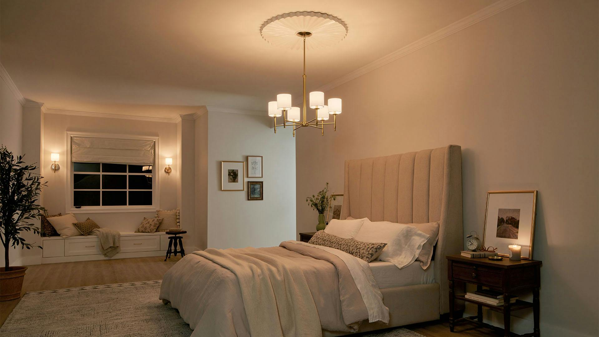 Bedroom at night with Ali 6-light chandelier and Ali wall sconces in brushed natural brass, lights turned on