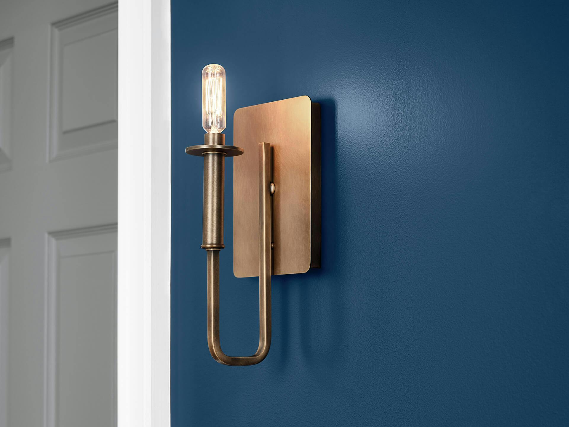 Close up of a brass alden sconce with light bulb on a blue painted wall