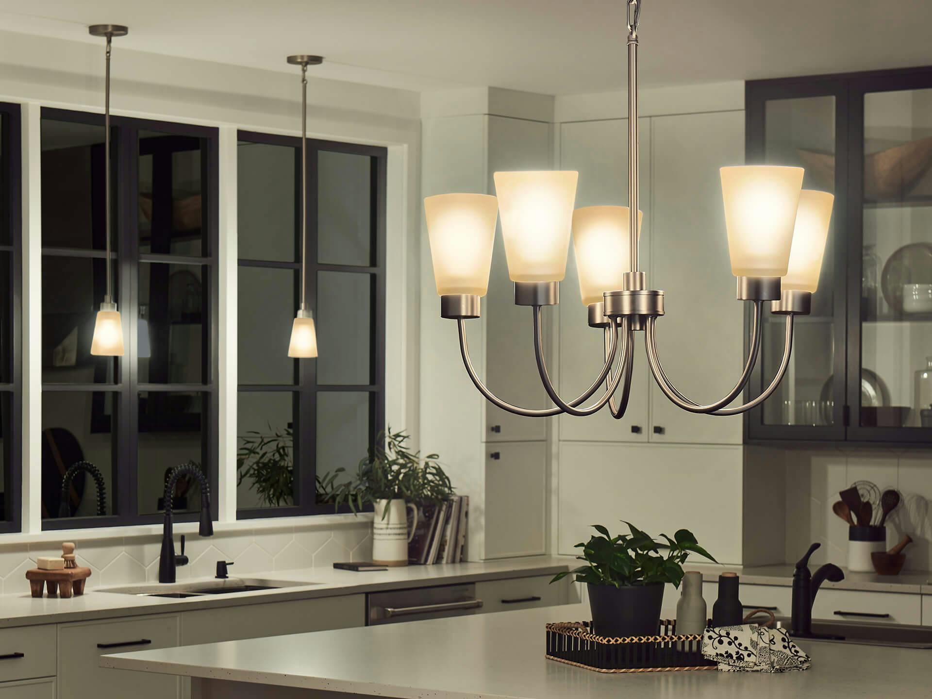 Modern kitchen with Emra chandelier and pendant lights over kitchen sink at night in brushed nickel finish