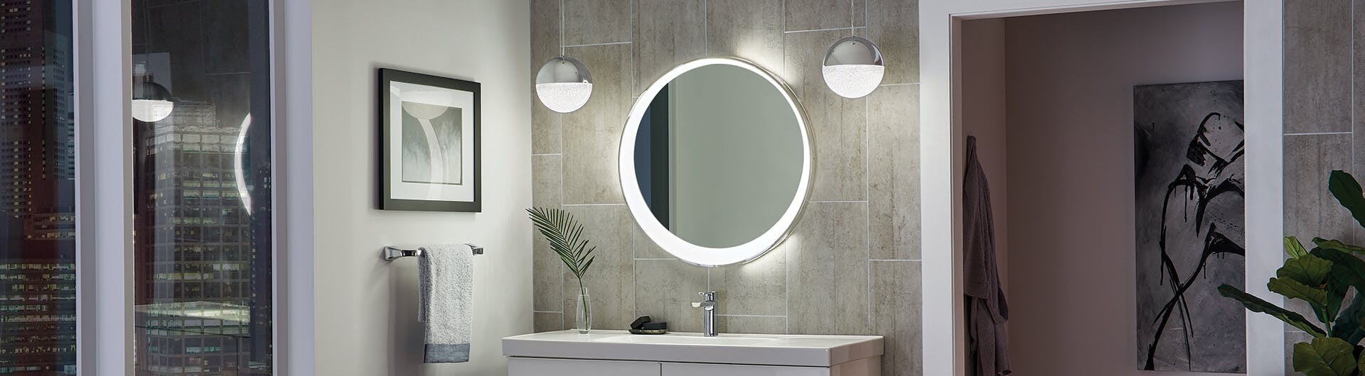 A bathroom at night with an offset round lighted mirror and two chome Moonlit pendants glowing in the center of the room