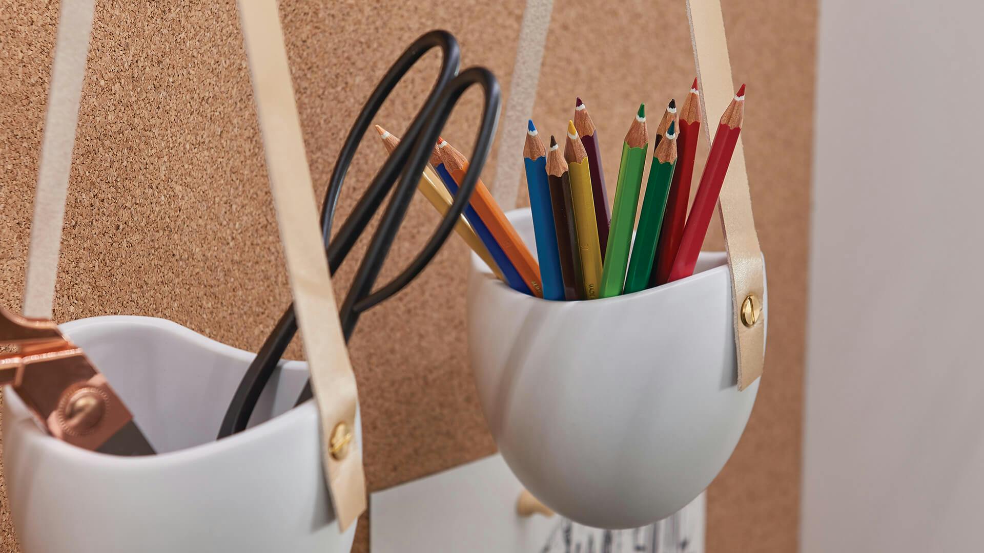 Pencils and scissors in hanging cups against a cork board with a drawing on it